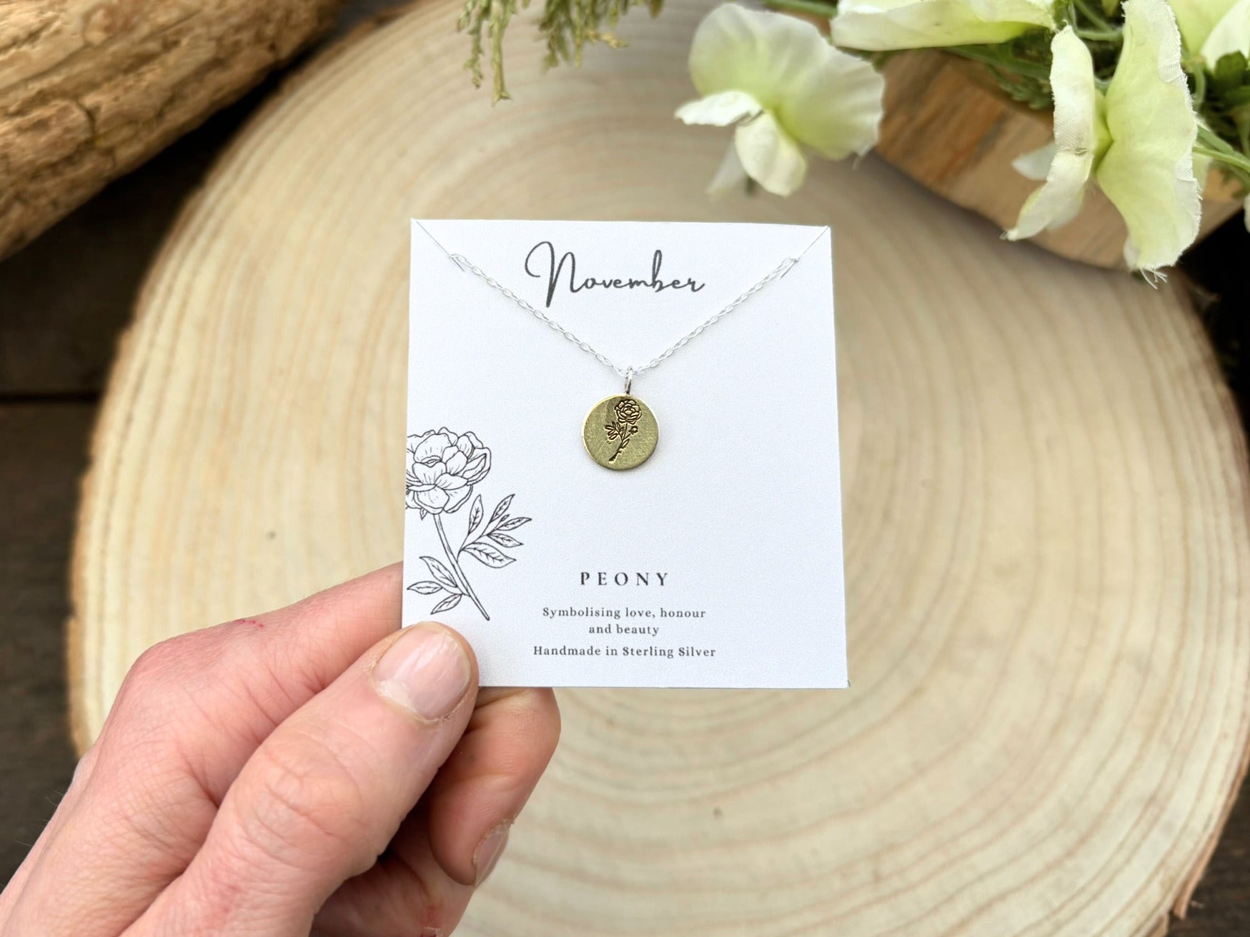 Necklace with a card labeled 'November' held by a hand on a wooden surface with flowers.