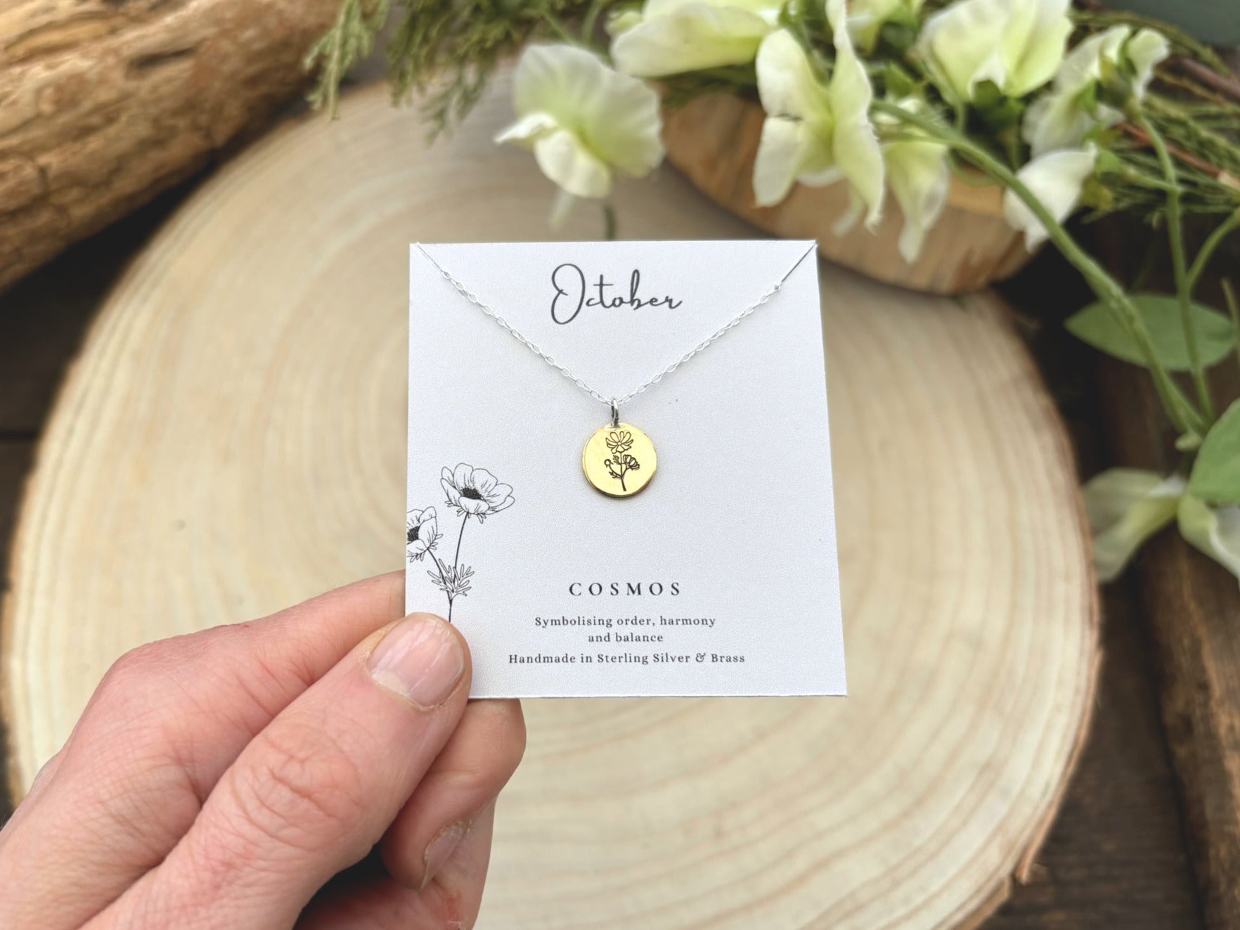 Gold necklace on a card held by a hand with a wooden surface and flowers in the background