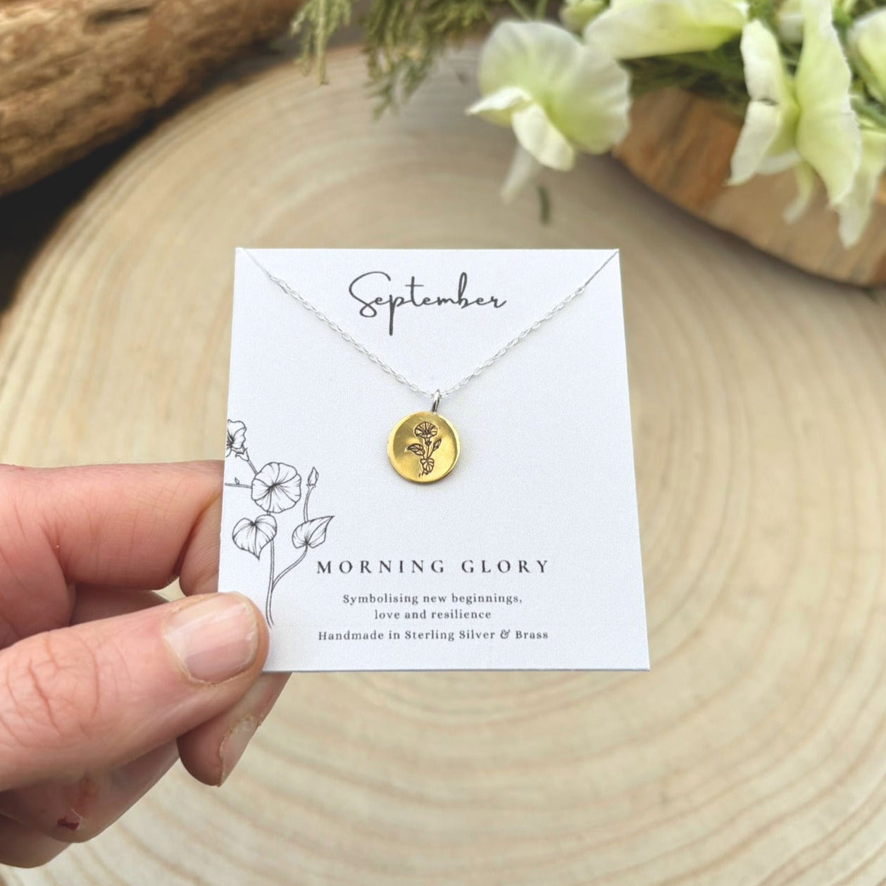 Hand holding a gold necklace with a card labeled 'September Morning Glory' on a wooden surface.