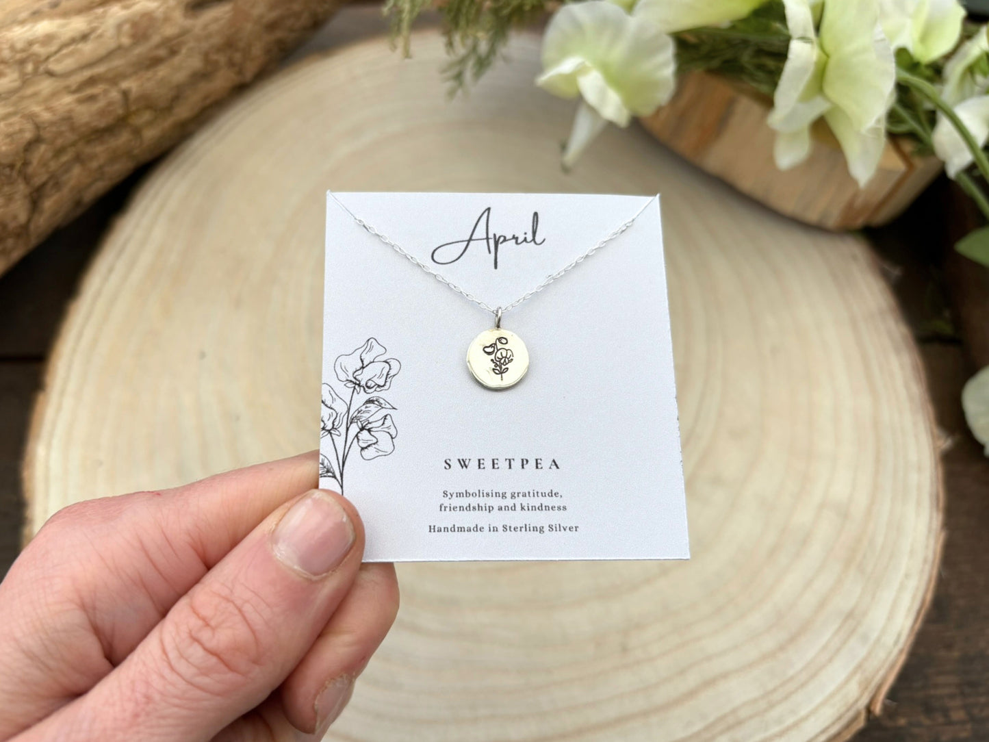 Necklace with a pendant on a card held by a hand, with flowers in the background, April birth flower, sweet pea