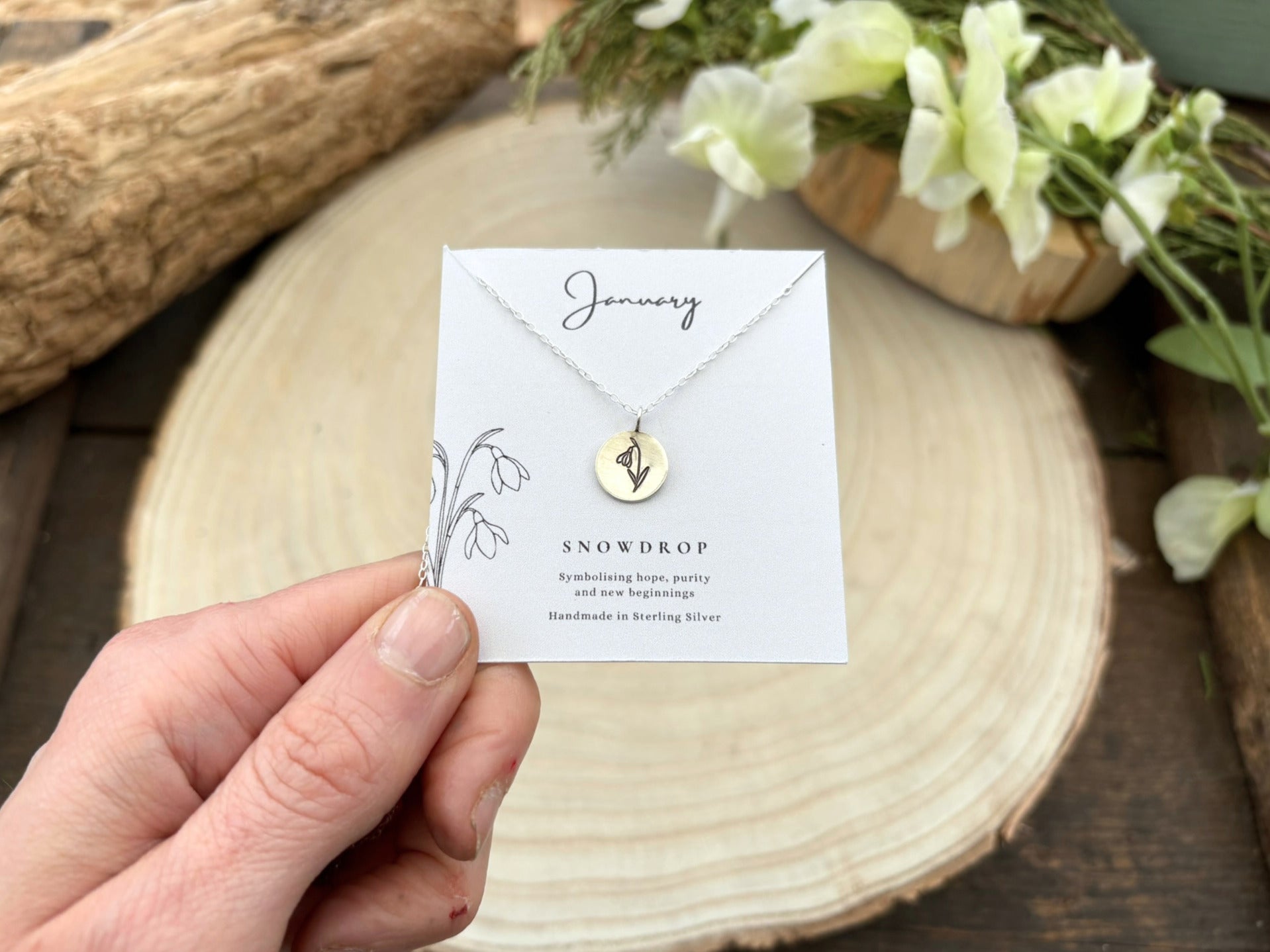 Hand holding a necklace card with a snowdrop design on a wooden surface with flowers. Birth flower necklace by Curious Magpie Jewellery