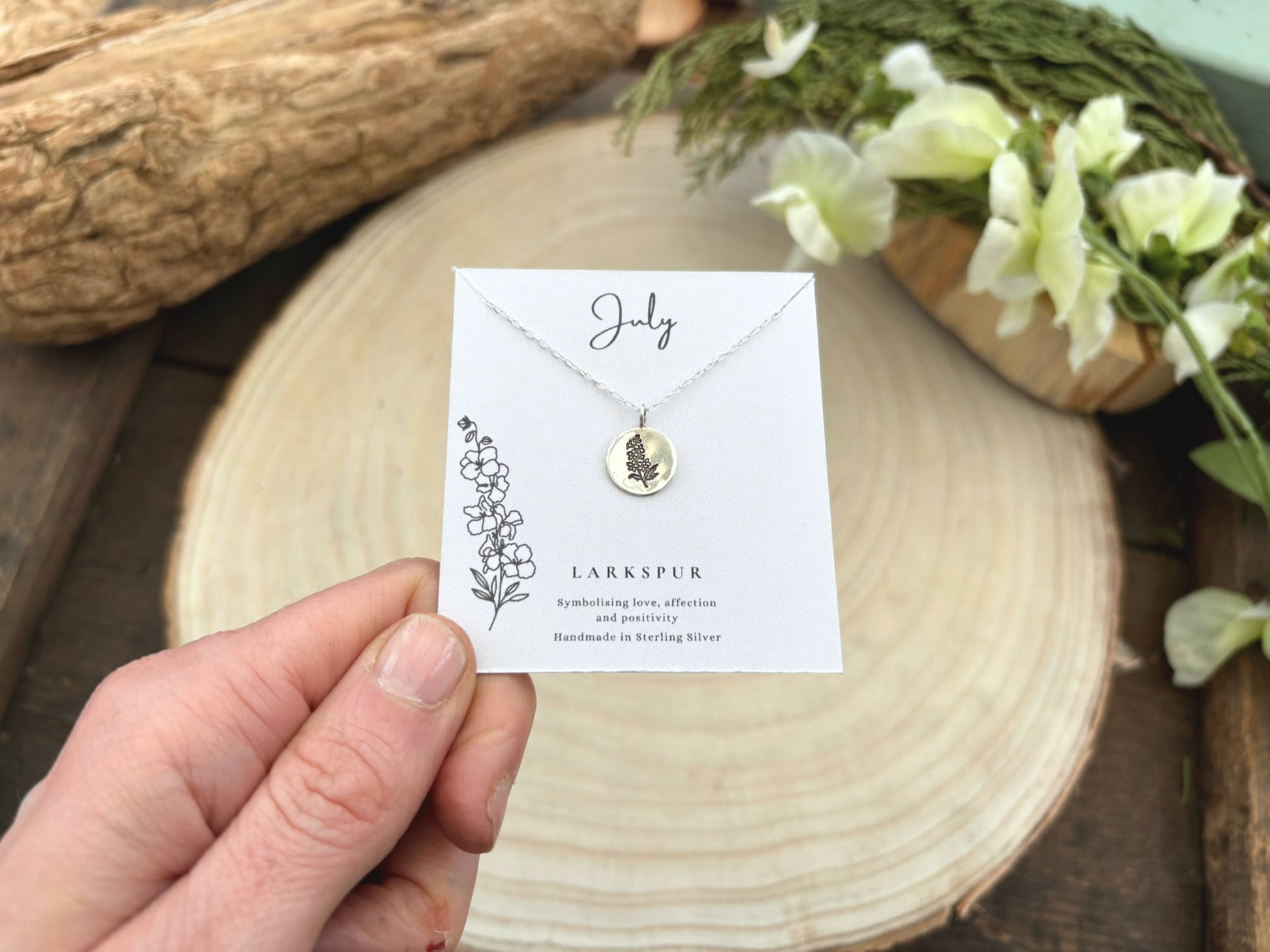Necklace with a card displaying 'July' and 'Larkspur' on a wooden surface with flowers.