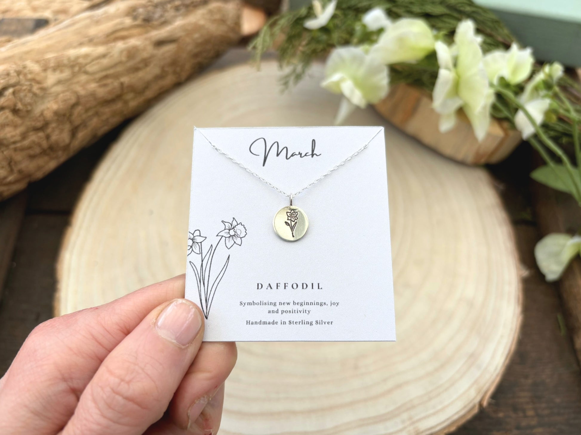 Necklace with daffodil charm on a card held by a hand, with flowers and wood in the background