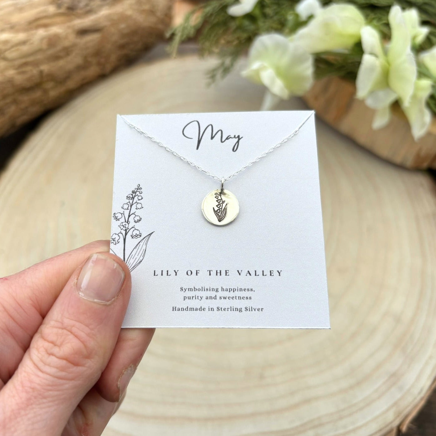 Hand holding a necklace card with a lily of the valley necklace on a wooden surface.