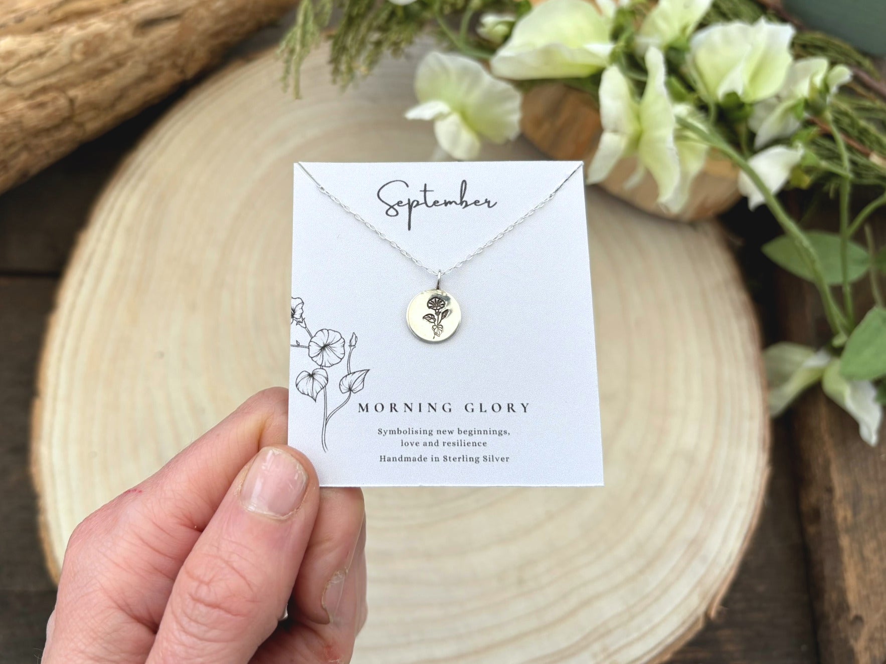 Necklace with a card labeled 'September Morning Glory' held by a hand on a wooden surface with flowers.